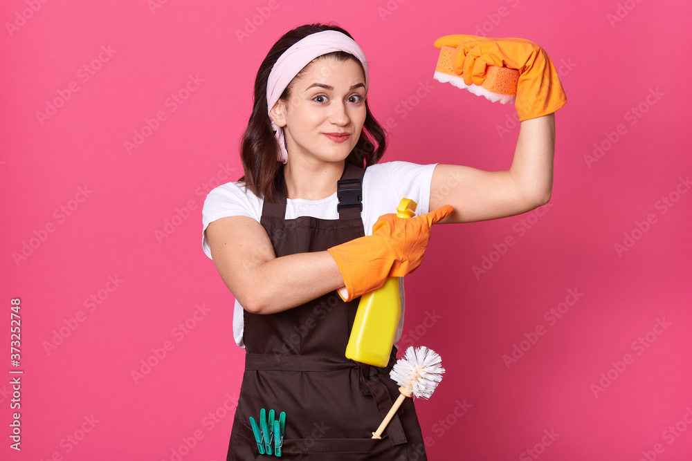 dark haired woman showing showing muscles isolated over rosy background, housekeeper wearing white t shirt and brown apron, holding sponge in hand. dark haired woman showing showing muscles isolated over rosy background, housekeeper wearing white t shirt and brown apron, holding sponge in hand.