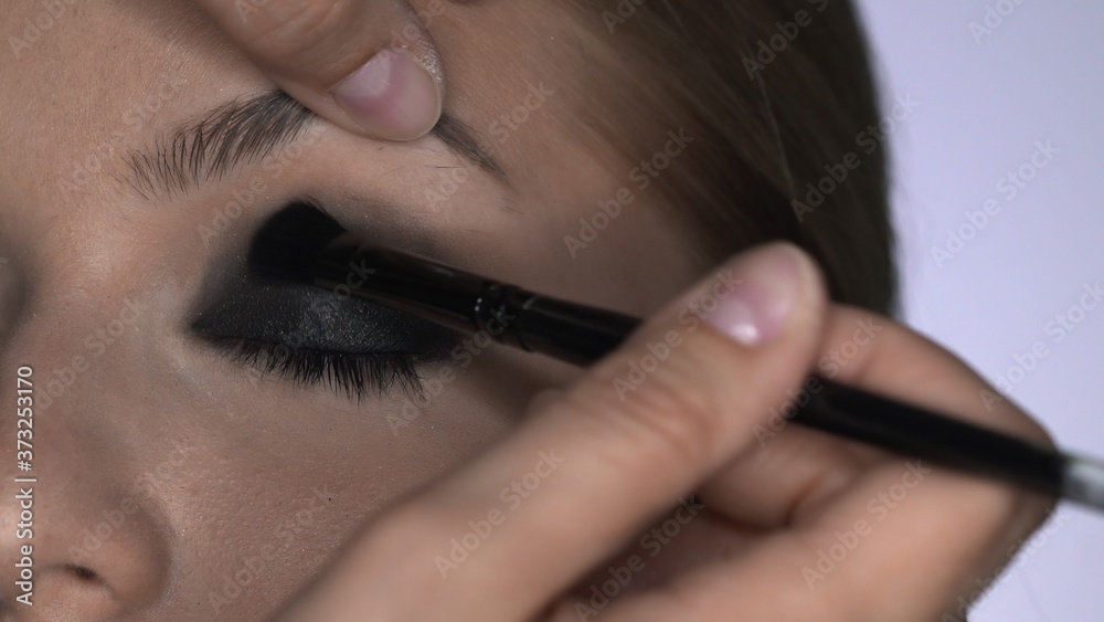 Makeup artist making professional make-up for young woman in beauty studio. Make up Artist makes the makeup smokey eyes of a black shade with special brush of a beautiful model