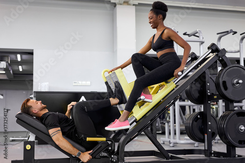 athletic people support each other in gym, do exercises together. man pumping legs on equipment with weight, african woman sit near, smile