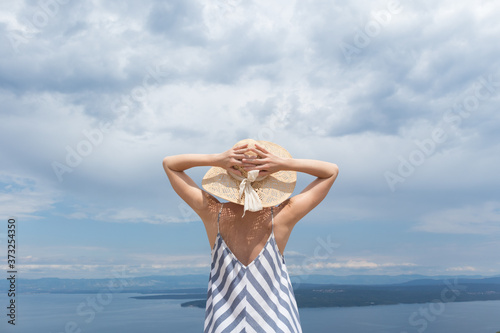 Rear view of young woman wearing striped summer dress and straw hat standing in super bloom of wildflowers, relaxing while enjoing beautiful view of Adriatic sea nature, Croatia. photo