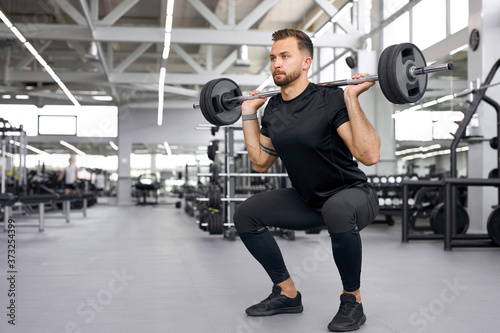 sportsman squats and raises a large barbell in gym, young strong guy do exercises, have muscular body