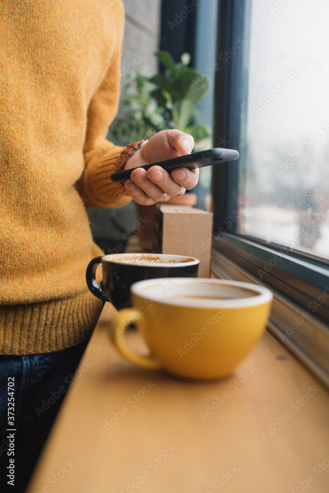 Two cups of coffee on wooden window sill and male hands holding cell phone on background. Lifestyle concept.