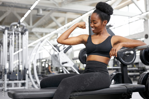 strong lady show arm muscles, sit in gym after training, happy woman in sportswear look at her arm and smile