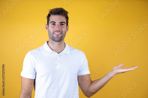 Young handsome man over isolated yellow background wearing casual t-shirt smiling to the camera while presenting with hand.