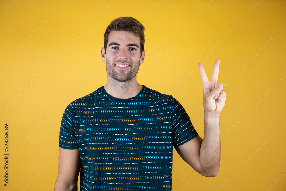 Young handsome man wearing casual blue t-shirt over yellow isolated background smiling with happy face winking at the camera doing victory sign. Number two. Young handsome man wearing casual blue t-shirt over yellow isolated background smiling with happy face winking at the camera doing victory sign. Number two.