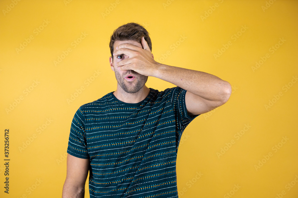 Young man standing over isolated yellow background peeking in shock covering face and eyes with hand, looking through fingers with embarrassed expression Young man standing over isolated yellow background peeking in shock covering face and eyes with hand, looking through fingers with embarrassed expression