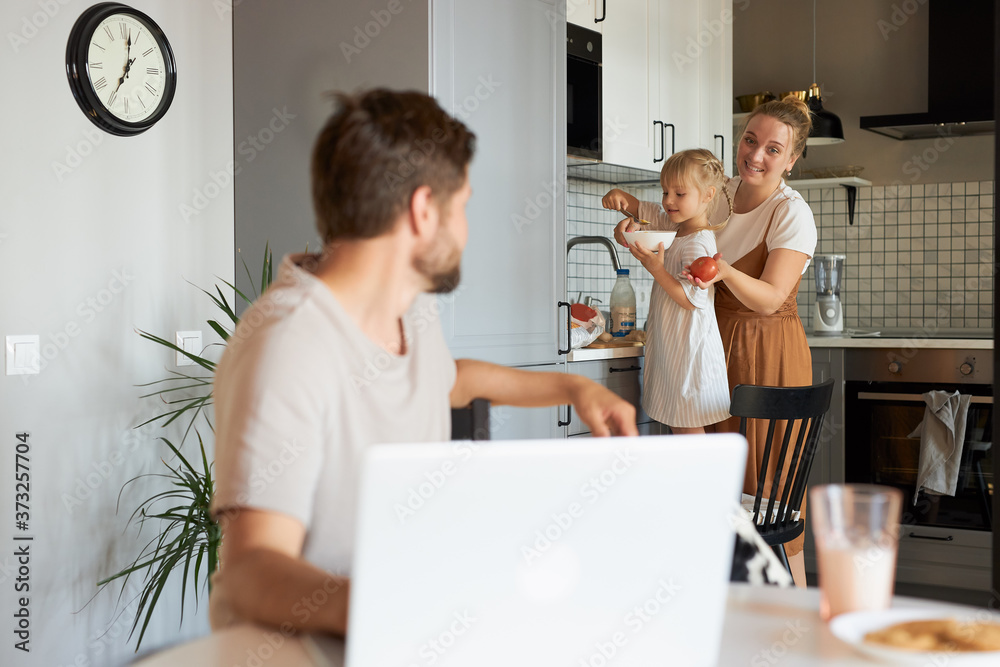adorable wife and daughter talk with working man in the kitchen, male sit using laptop. at home