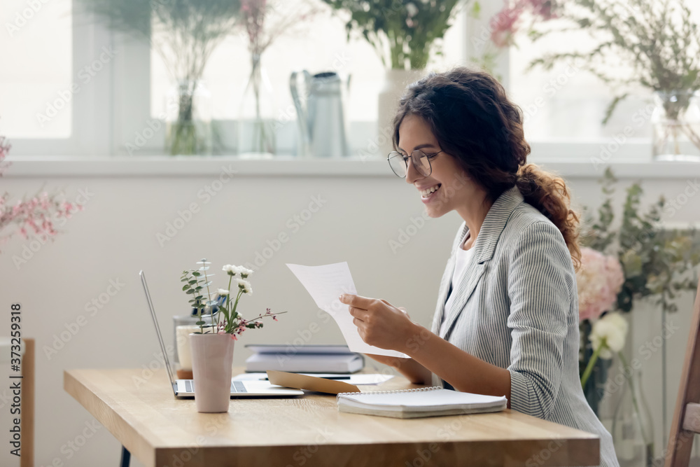Side view pleasant smiling young businesswoman creative designer in eyeglasses reading paper letter correspondence with good news or banking notification, sitting at desk in modern showroom office. Side view pleasant smiling young businesswoman creative designer in eyeglasses reading paper letter correspondence with good news or banking notification, sitting at desk in modern showroom office.