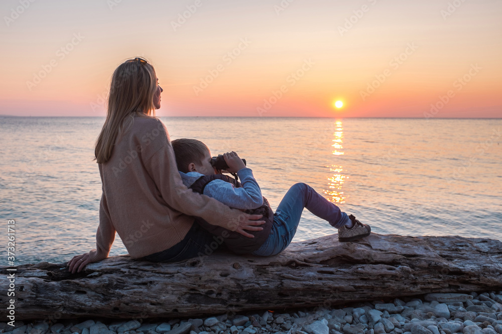 Mother and son tourist traveling sitting on the beach and look binocular under sunset sky with sun in summer. Mothers Day.