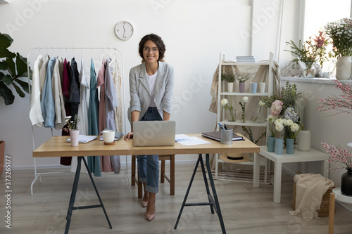 Full length portrait of attractive smiling young stylish fashion clothes designer in eyeglasses standing near table with computer, enjoying working in modern showroom atelier, casual workday concept.