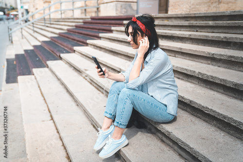 Beautiful young caucasian woman looking at her smartphone and smiling in urban background. Brunette girl wearing casual clothes sitting on stairs.