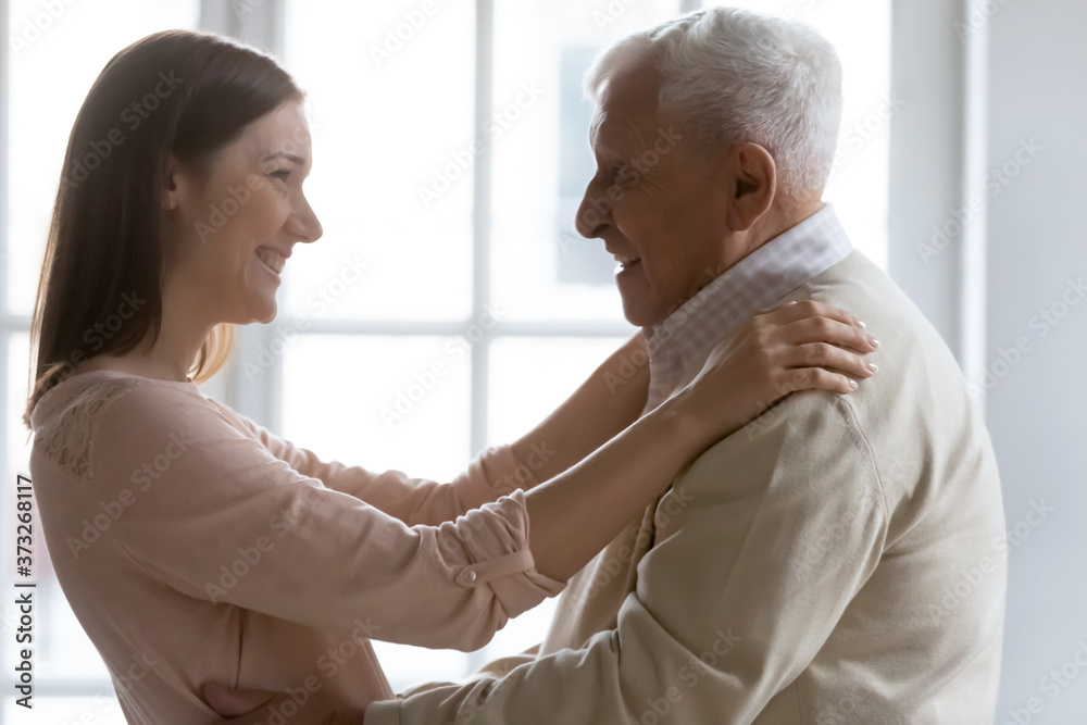Obraz premium Side view happy positive elderly senior man greeting grown up child daughter at home. Smiling beautiful young woman glad to see older 70s father, enjoying visit at weekend free leisure time.