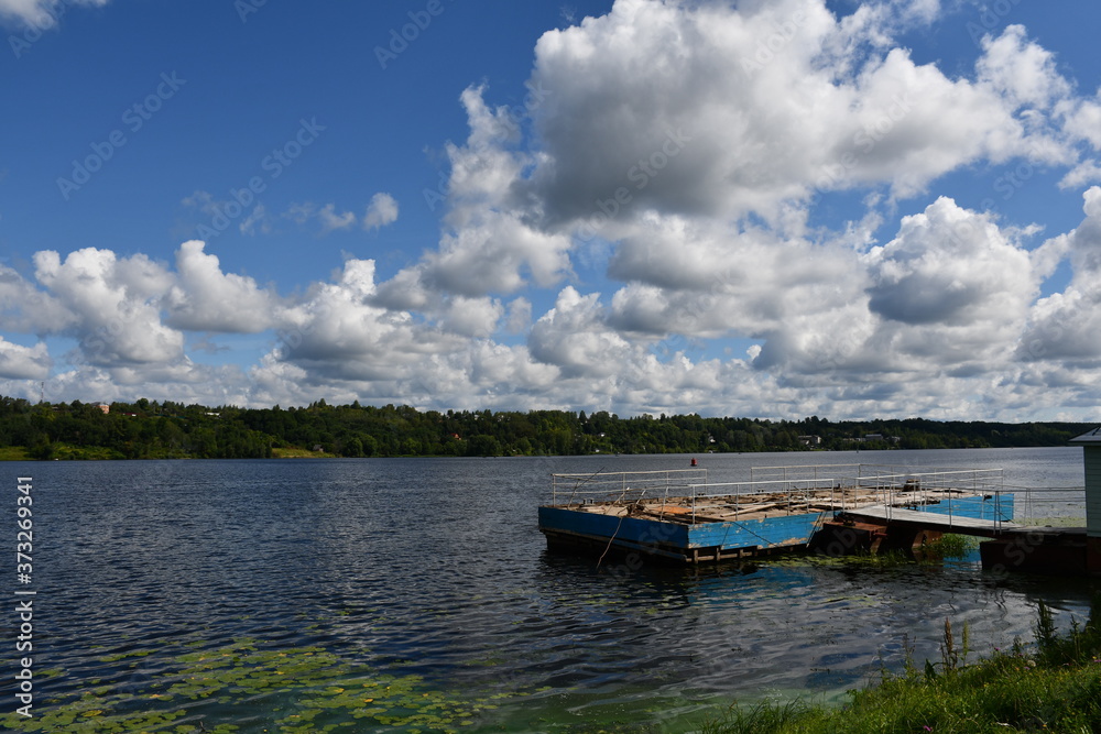 Panorama of a large river or lake. Water lilies grow near the shore. A small old dilapidated pier for boats and motorboats. Beautiful clouds.