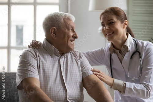 Smiling kind young nurse cuddling shoulders of elderly mature retired male patient, supporting indoors. Happy old 80s man enjoying conversation with pleasant doctor, medical insurance concept.