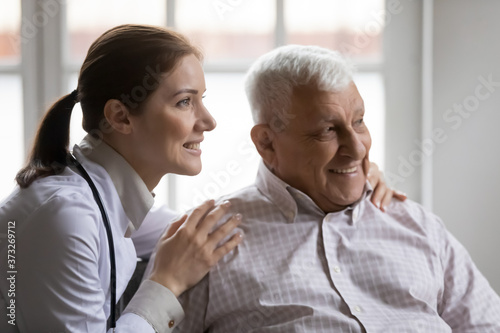 Dreamy young female doctor cuddling shoulders of smiling old retired patient  looking together at distance. Head shot happy caring nurse supporting elderly mature man  thinking of healthy lifestyle.