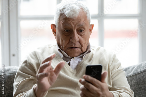 Shocked 70s retired old man looking at smartphone screen, reading message with unexpected news at home. Surprised elderly grandfather having problems with using mobile applications, tech concept. photo