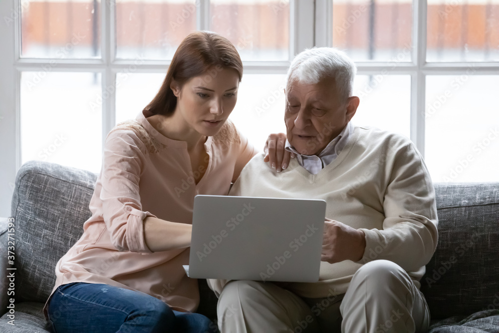 Focused young woman helping older daddy with computer applications, sitting together on comfortable sofa in living room. Serious different generations enjoying web surfing information in internet.