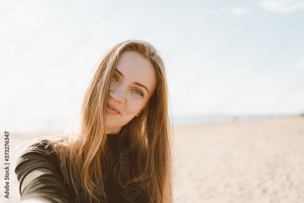 Pretty female with long hair, blonde takes selfie photo on mobile phone. Beautiful woman looking at camera and tilting head. Sandy beach in unny day in ocean or sea coastline