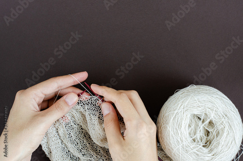 female hands knitting scarf with white lace yarn on wood knitting needles. dark background, top view