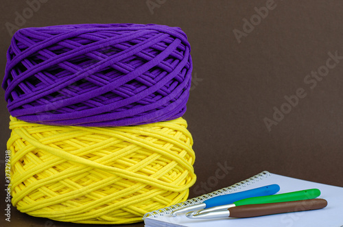 yellow and purple t-shirt yarn balls and colorfull crochet hooks on notebook on brown background. Craft, handmade, hobby concept.