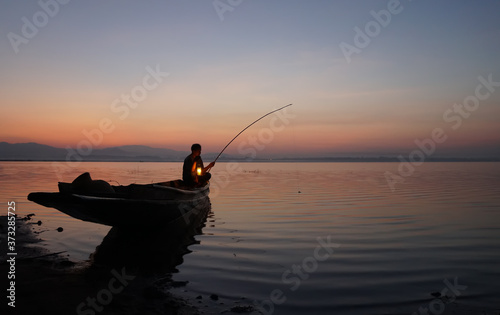 At lake side, asian fisherman sitting on boat and using fishing rod to catch fish at the sunrise