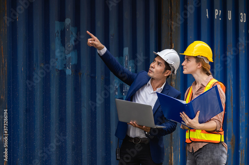 two engineer worker hold a laptop, document for checking a quality of containers box from cargo ship for export and import, blue container background