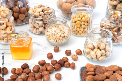 An assortment of different nuts in glass jars: hazelnuts, cashews, pistachios, pine nuts, walnuts, almonds and chickpeas. Jars of nuts are on a white table, near a small jar of honey