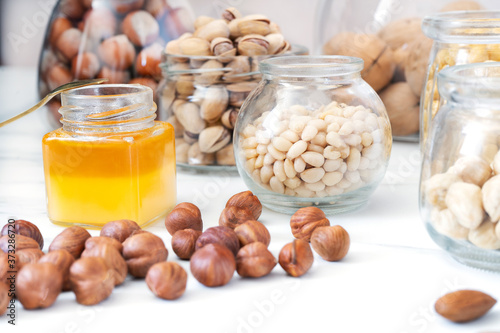 Peeled hazelnuts are scattered on a white table. There are jars of honey and nuts (pine nuts, pistachios, hazelnuts in shells, walnuts) nearby. Raw food concept. Vegetarian food. Close-up