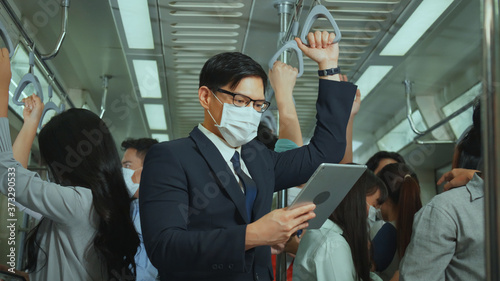 Asian businessman wear mask while traveling to work on skytrain. Prevent Coronavirus, bacteria in public place from people. Business people use tablet outside meeting at peak hour no social distancing