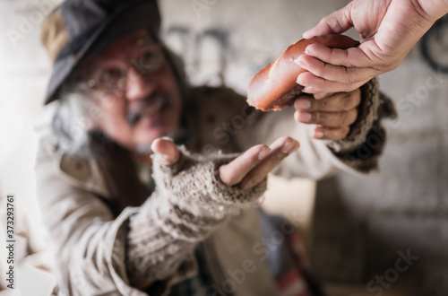 Old homeless man with gray beard sitting hungry and food beggar from people walking on street. Closeup and focus to her hand with giving bread for poor old man homeless. Sharing and hope concept.