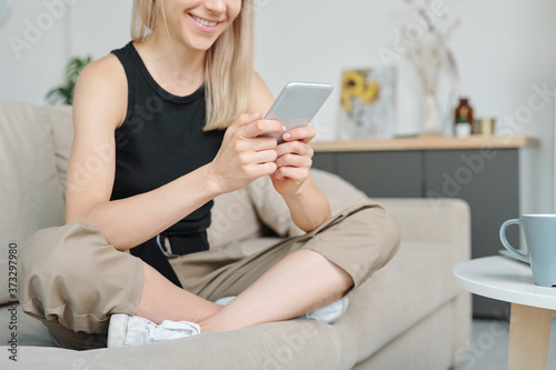 Happy female millennial sitting on couch with her legs crossed and texting