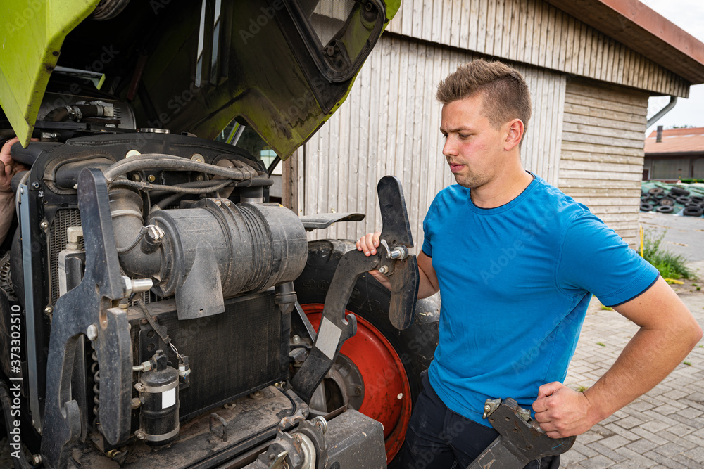 Fototapeta premium Landwirtschaft und Landtechnik, junger Landwirt vor geöffneter Motorhaube eines Traktors