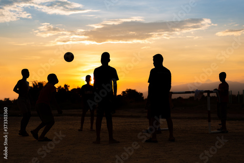 Silhouette action sport outdoors of a group of kids having fun playing soccer football for exercise in community rural area under the twilight sunset. Poor and poverty children in development country.