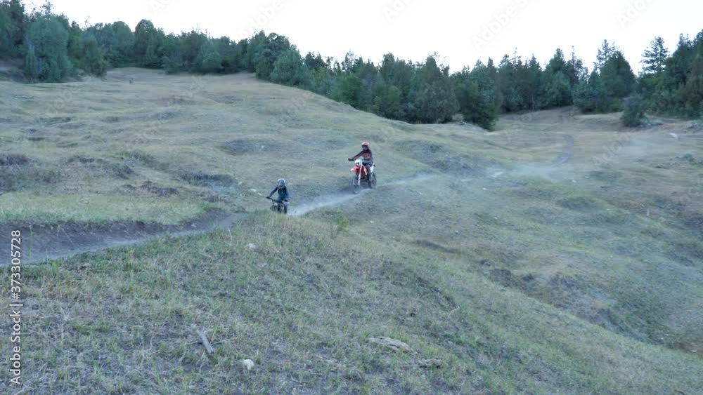 Son rides a motorcycle along a dusty trail with father following closely behind - slow motion Son rides a motorcycle along a dusty trail with father following closely behind - slow motion