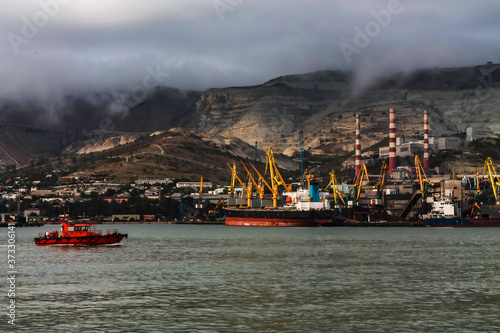 A red harbor tug sailing in the bay against the background of a cargo ship moored in the seaport against the background of mountains and factory pipes. Cloudy weather in the mountains. © Алекс Швачко