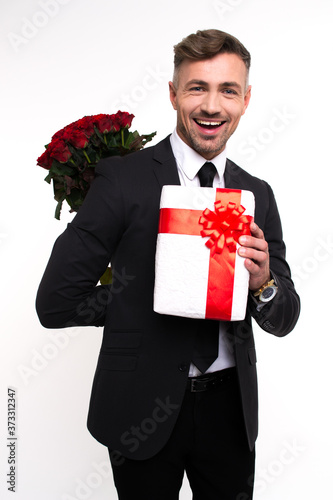 Hello my love! Handsome young man holding roses behind his back and looking at camera with smile while standing against white background 