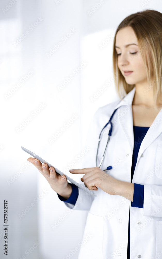 Young woman-doctor is holding a tablet computer in her hands, while standing in a clinic. Portrait of friendly female physician with a stethoscope at work. Perfect medical service in a hospital Young woman-doctor is holding a tablet computer in her hands, while standing in a clinic. Portrait of friendly female physician with a stethoscope at work. Perfect medical service in a hospital