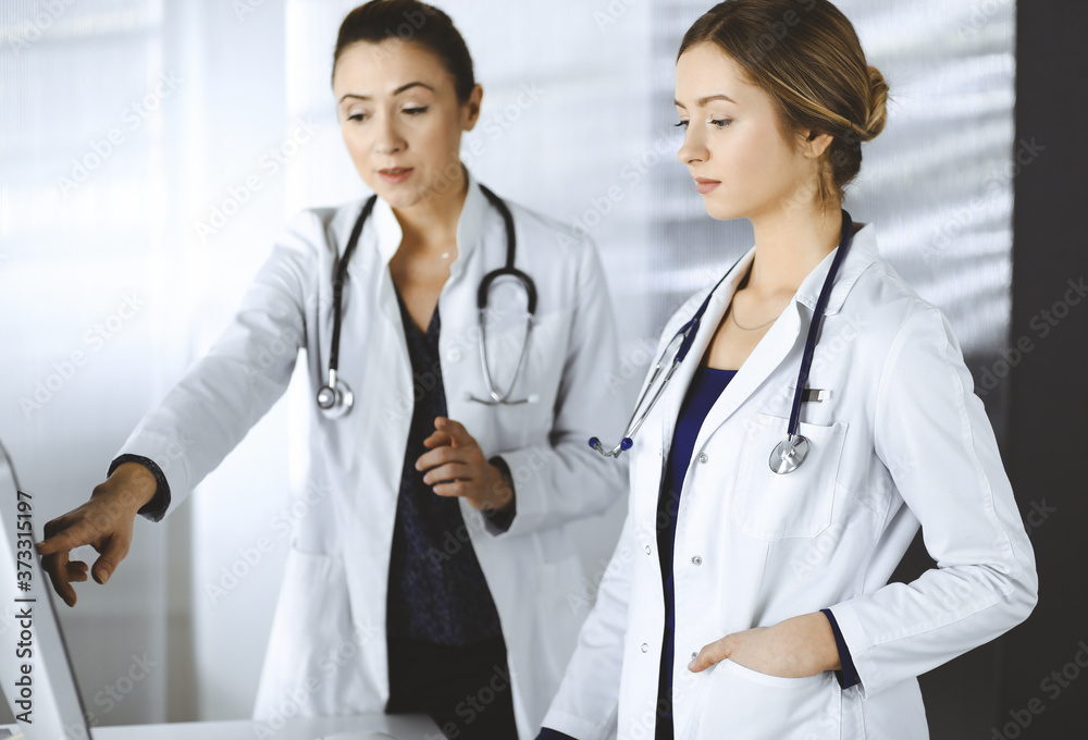 Two female physicians are discussing medical therapy, while standing at the table in a clinic office. Doctors use pc computer at work. Teamwork in medicine Two female physicians are discussing medical therapy, while standing at the table in a clinic office. Doctors use pc computer at work. Teamwork in medicine