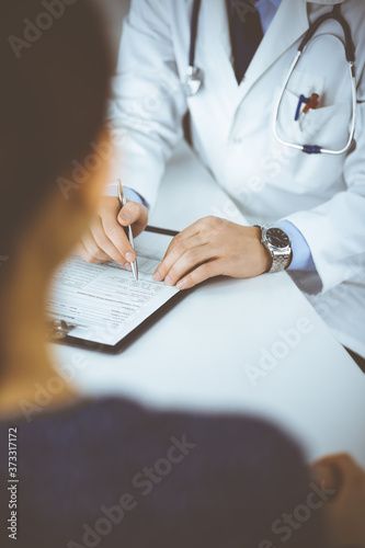 A doctor is talking to his patient, while sitting together at the desk in the cabinet in a hospital. Physician using clipboard for filling up medication history records. Perfect medical service in