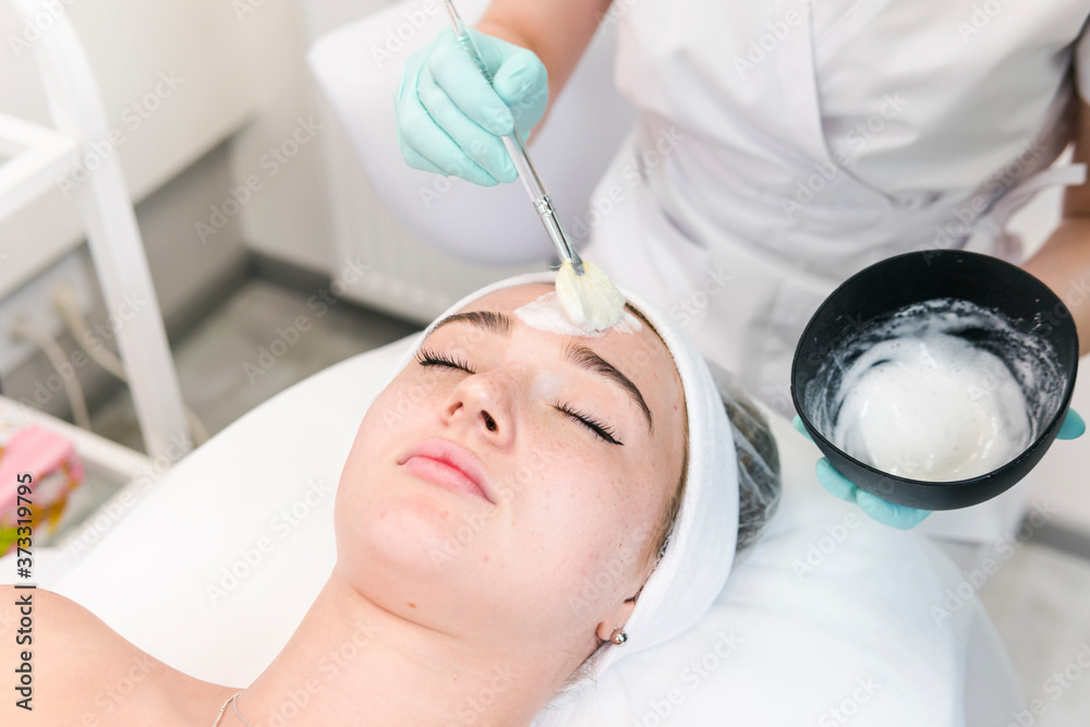 Naklejka premium The young female client of cosmetic salon having a cleansing facial mask. The procedure of applying a peeling mask to the face. Concepts of skin care and beauty salon or clinic.