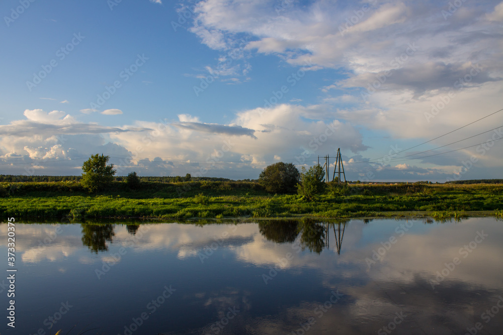 Pastoral landscape-Klyazma river Bank with green trees and grass against a blue cloudy sky with reflections in the water and space for copying