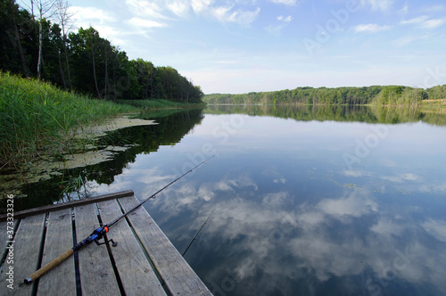 fishing rod on a wooden pier and reflection of clouds in the lake water