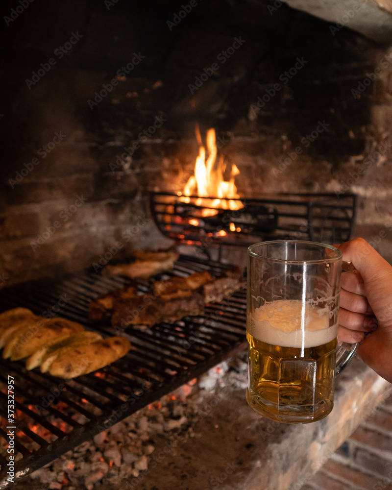 Carne y verduras cocinandose a las brasas, y una mano sostiene una cerveza.