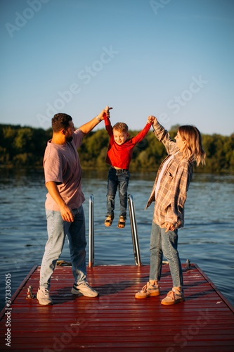 Happy young family raise the baby boy by the hands up on river background. Summer photography for blog about family