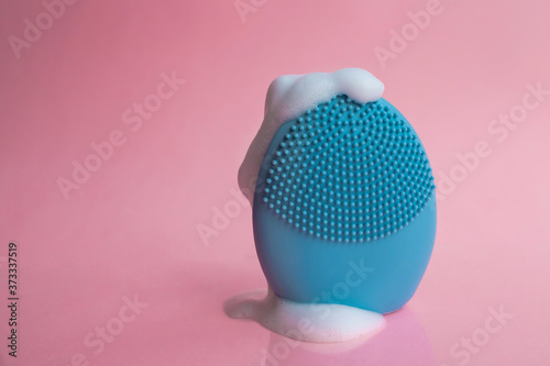 Silicone brush for washing face in foam. Blue facial cleansing brush on pink background. Beauty and skincare concept. Brush for face lifting, anti-aging wrinkles and massage