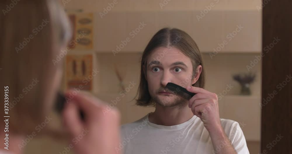 Young man looks at himself in the bathroom mirror and combing his mustache Young man looks at himself in the bathroom mirror and combing his mustache