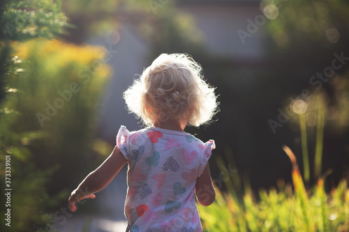 Backview of happy cheerful small girl with blond curly hair running in the garden in sunny summer day