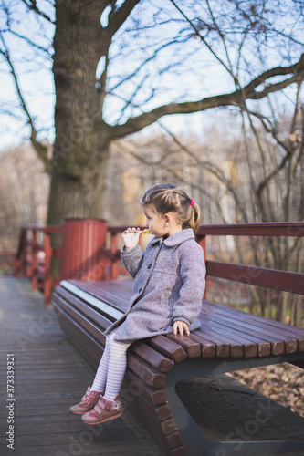 Elegant stylish little girl with pigtails in trendy grey coat, tights, snakers eats lollipop seating on wooden bench in fall park outdoors. Fashion autumn photo shoot. Family walk. Profile side view