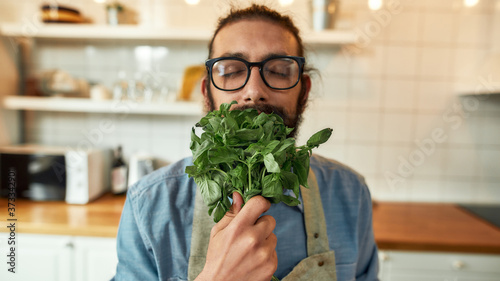 Photography Close up of young man, Italian cook in apron smelling basil leaves while getting ready to prepare a meal, standing in the kitchen