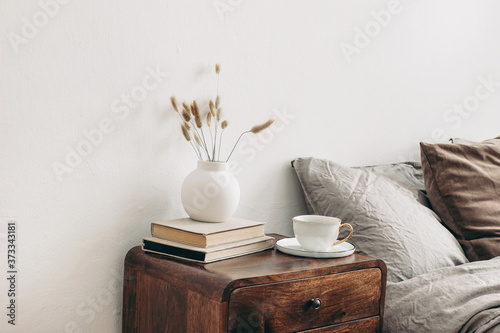 Modern white ceramic vase with dry Lagurus ovatus grass and cup of coffee on retro wooden bedside table. Beige linen and velvet pillows in bedroom. Scandinavian interior. Homestaging. photo
